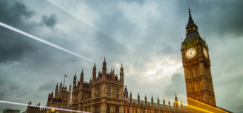 Big Ben and the Houses of Parliament in London, England. The building is lit up and set against a dramatic sky with cars and busses zooming by on Waterloo Bridge.