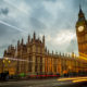 Big Ben and the Houses of Parliament in London, England. The building is lit up and set against a dramatic sky with cars and busses zooming by on Waterloo Bridge.